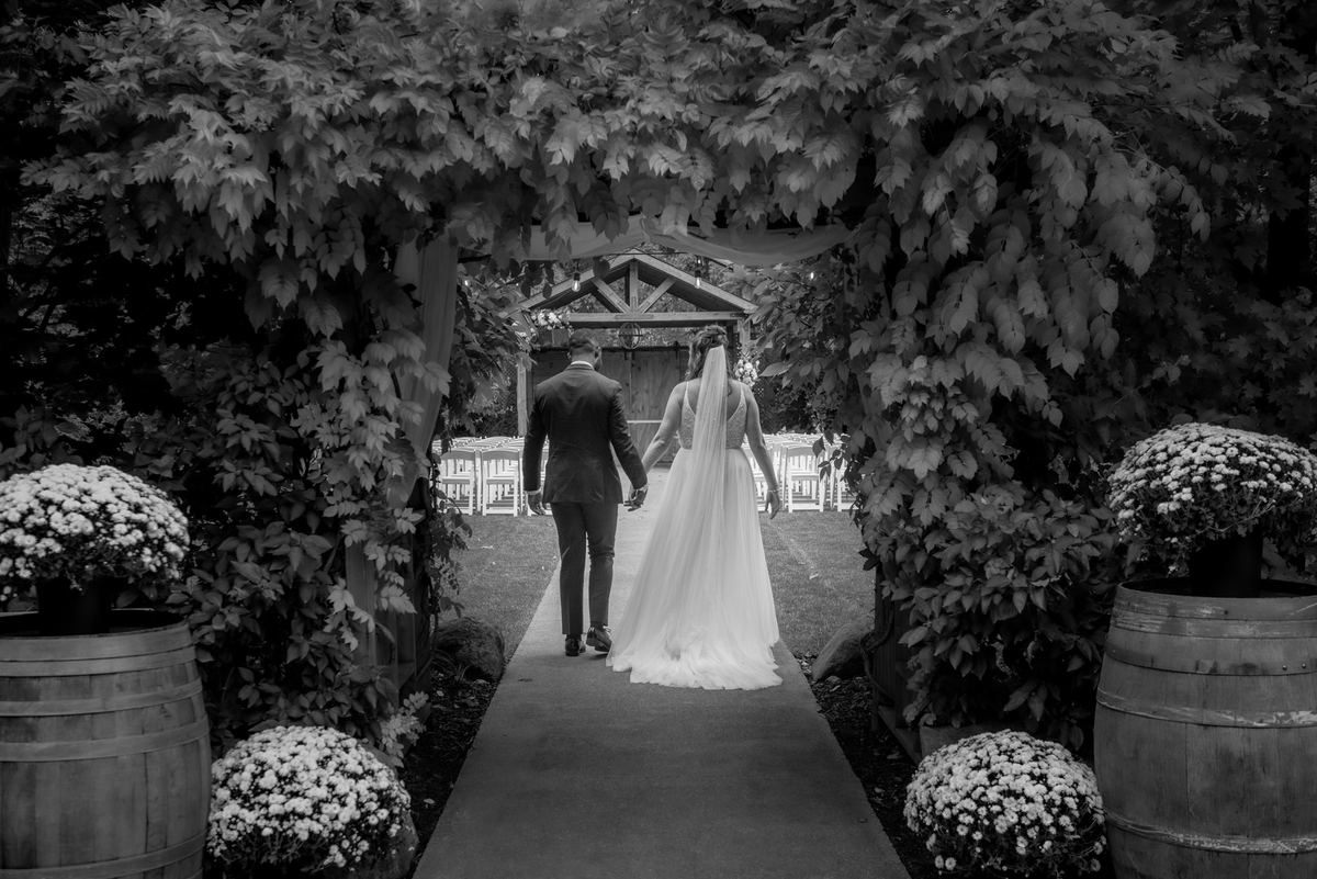A bride and groom walk down an aisle framed by lush greenery and white flowers, approaching an outdoor ceremony arch.