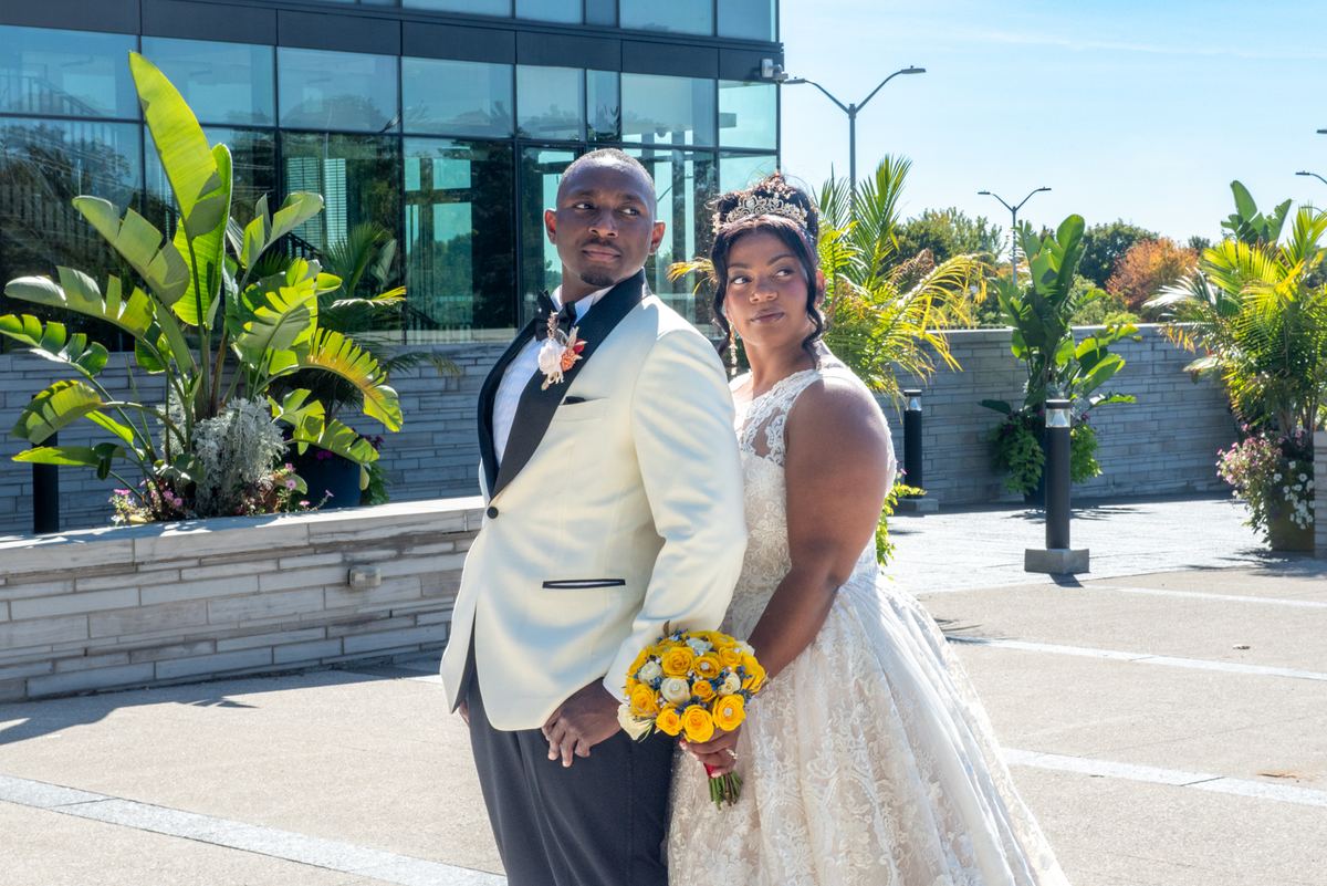 A newlywed couple in formal attire stand outdoors with lush tropical foliage and a modern glass building behind them.