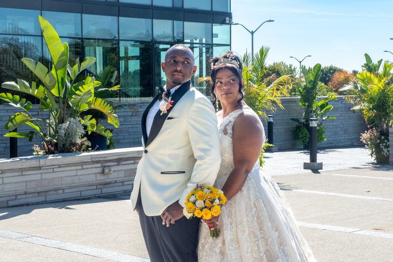 A newlywed couple in formal attire stand outdoors with lush tropical foliage and a modern glass building behind them.