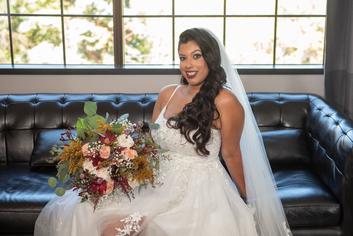 A bride in a wedding dress holds flowers and smiles on a sofa near a window, bathed in natural light.
