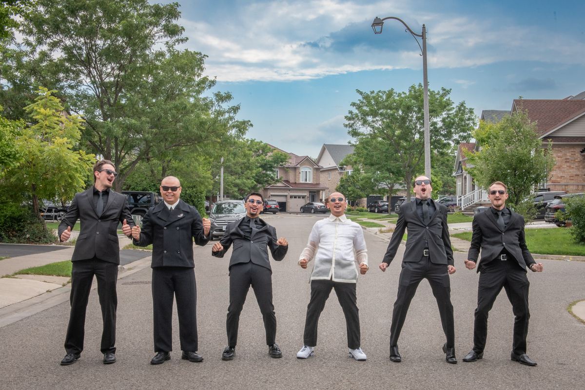 Six formally dressed men pose energetically on a suburban street, arms outstretched against a backdrop of houses and trees.