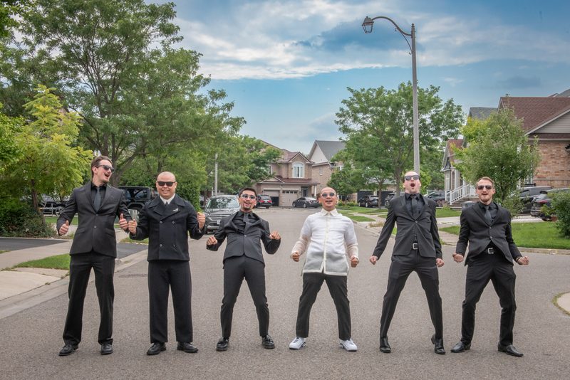 Six formally dressed men pose energetically on a suburban street, arms outstretched against a backdrop of houses and trees.
