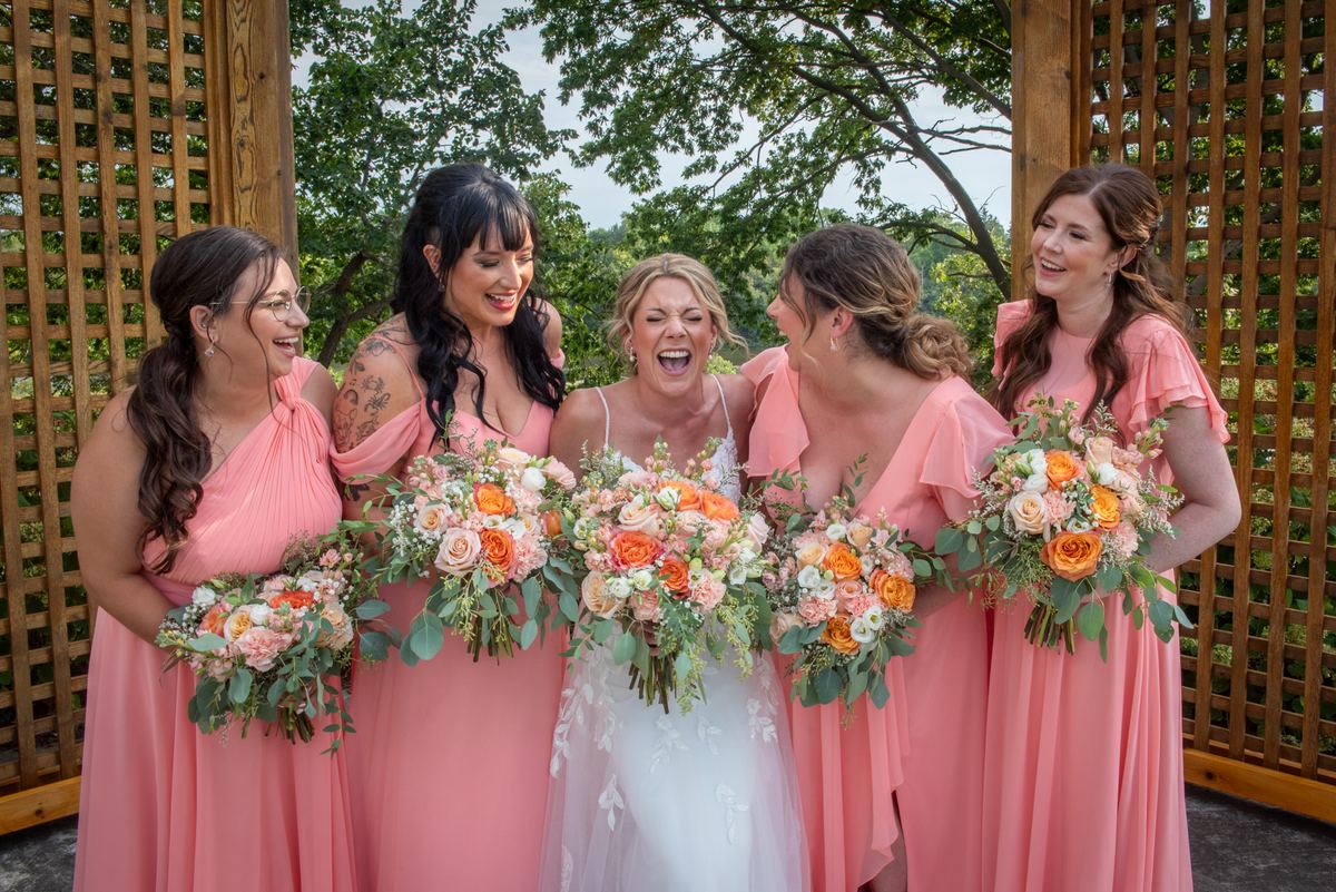 A bride and her bridesmaids laugh together under a wooden trellis surrounded by green foliage, holding their floral bouquets.