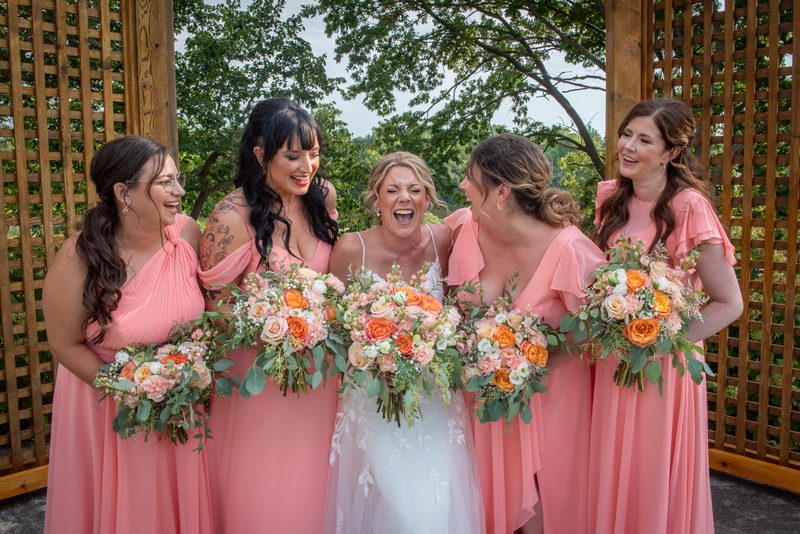 A bride and her bridesmaids laugh together under a wooden trellis surrounded by green foliage, holding their floral bouquets.