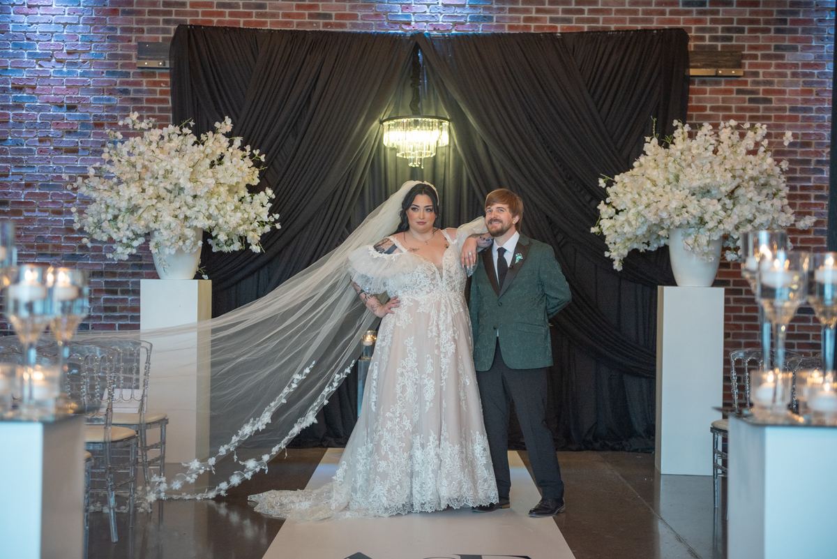 Bride in lace dress and veil with groom in green suit stand on white runner near floral arrangements.