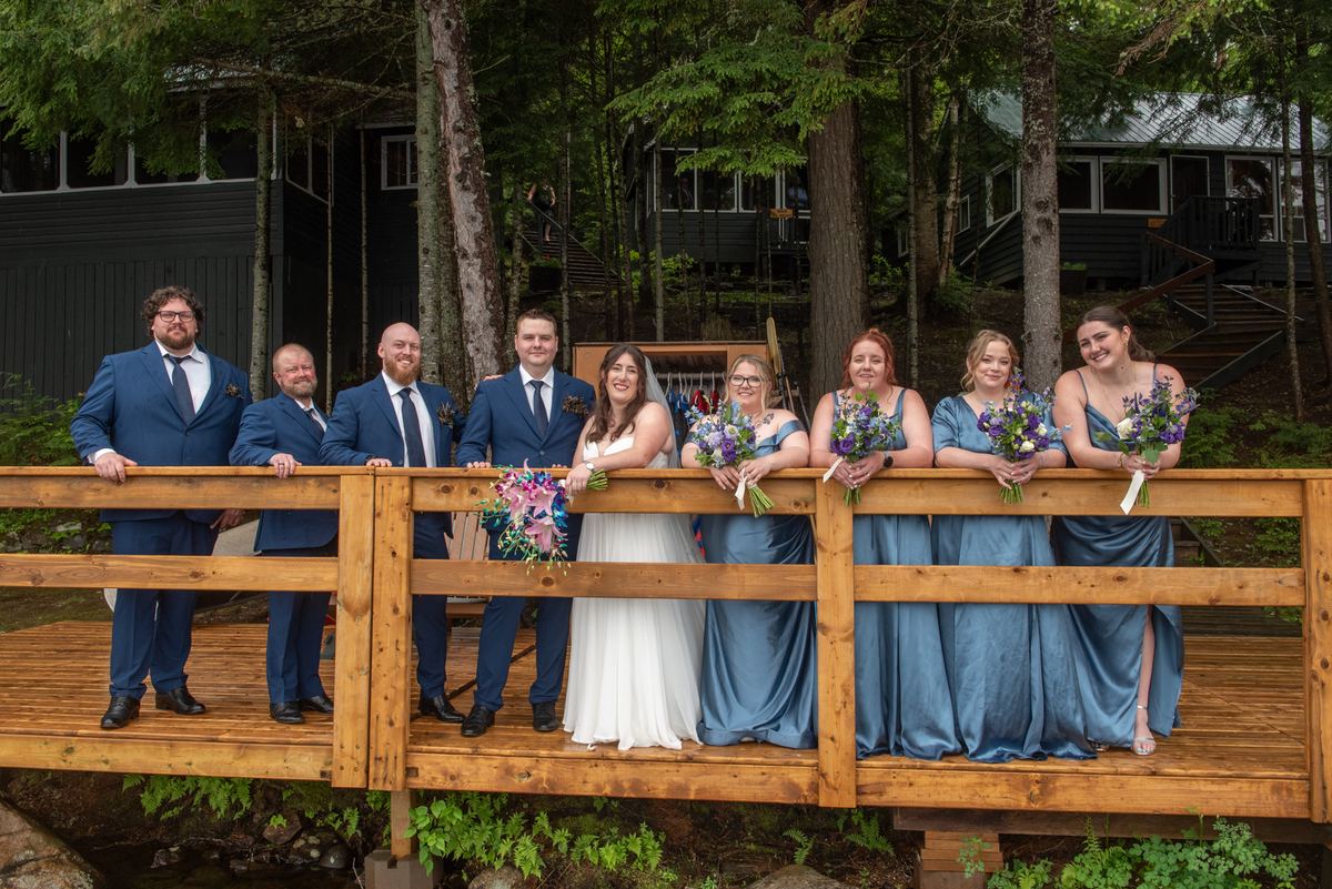 A wedding party poses on a rustic wooden bridge surrounded by dense forest in soft, diffused daylight.