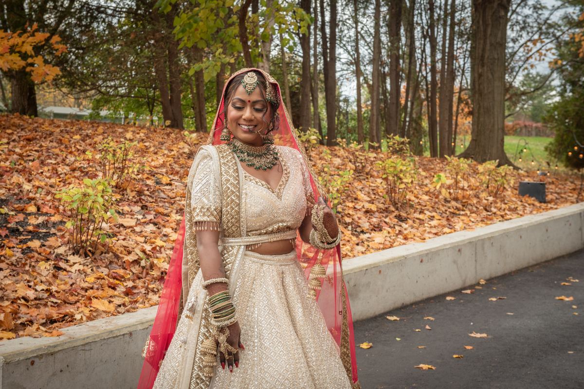 A smiling bride in a golden lehenga and red dupatta walks along an autumn path lined with leaves, surrounded by trees.