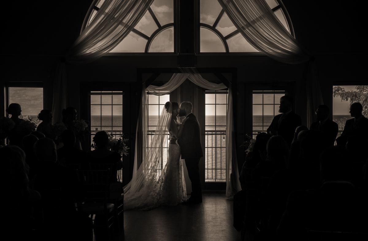 Bride and groom kiss at an altar, silhouetted against a sunset over water with guests in shadowed rows behind them.