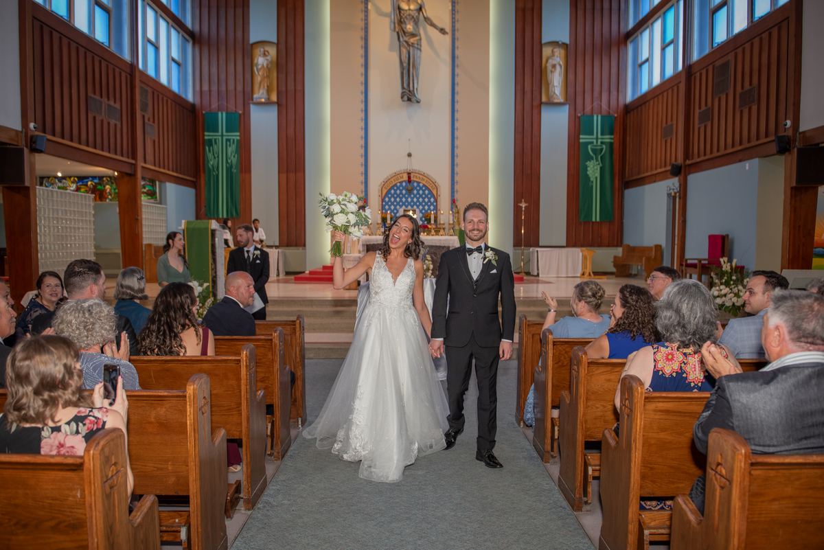 A smiling bride and groom exit the church aisle, hand in hand, as sunlight fills the space through tall windows.