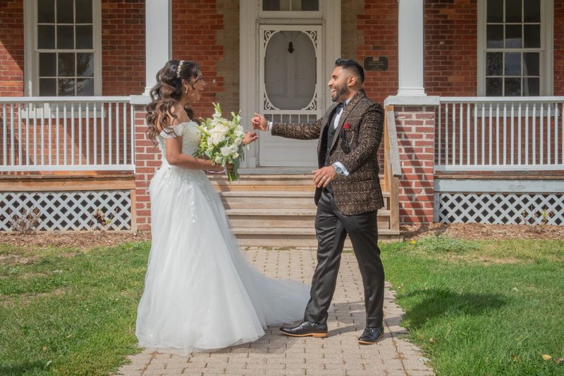 Fazana and Aman stand on a brick pathway before a red brick house, smiling and reaching toward each other.