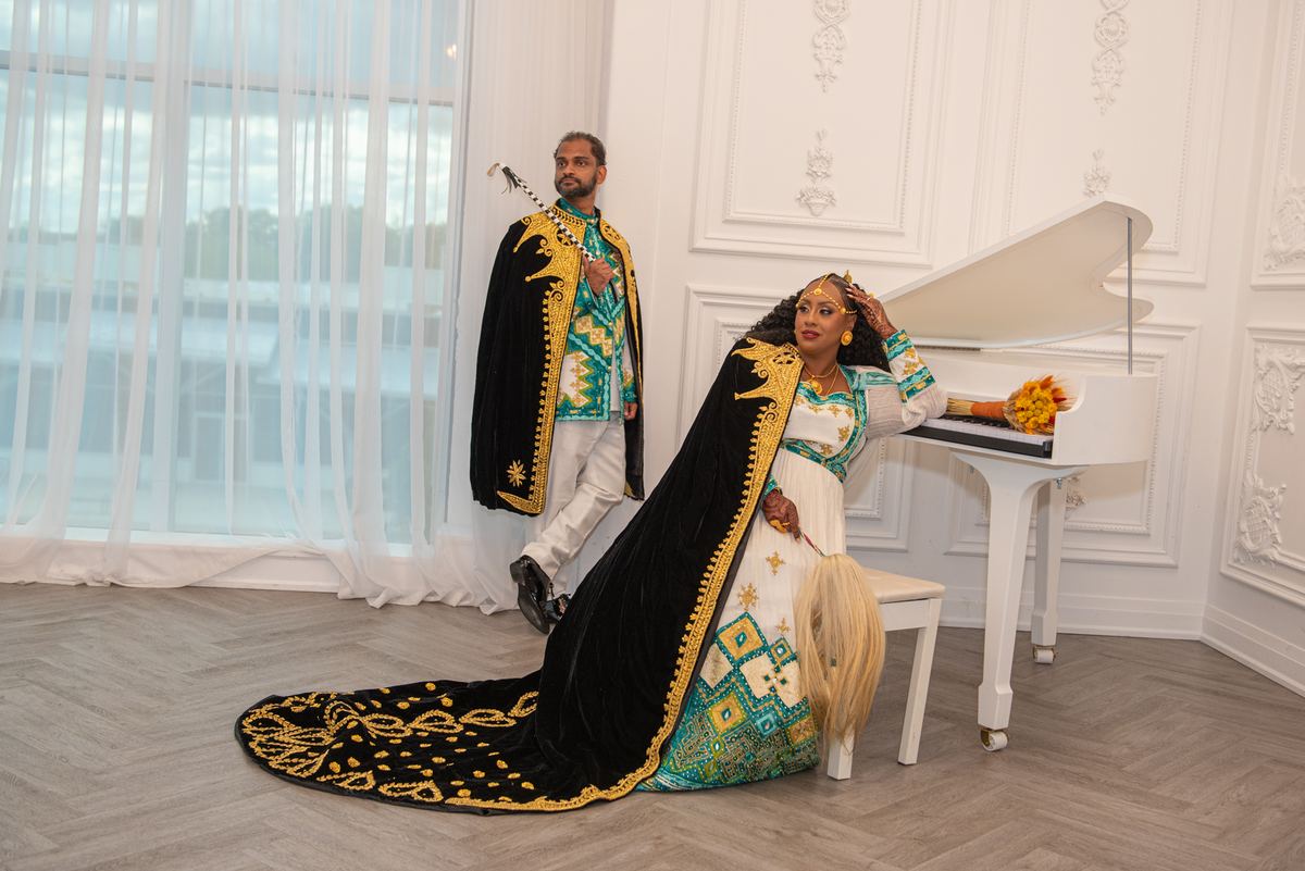 A man and woman in ornate traditional attire pose gracefully beside a grand piano in an elegantly appointed room.
