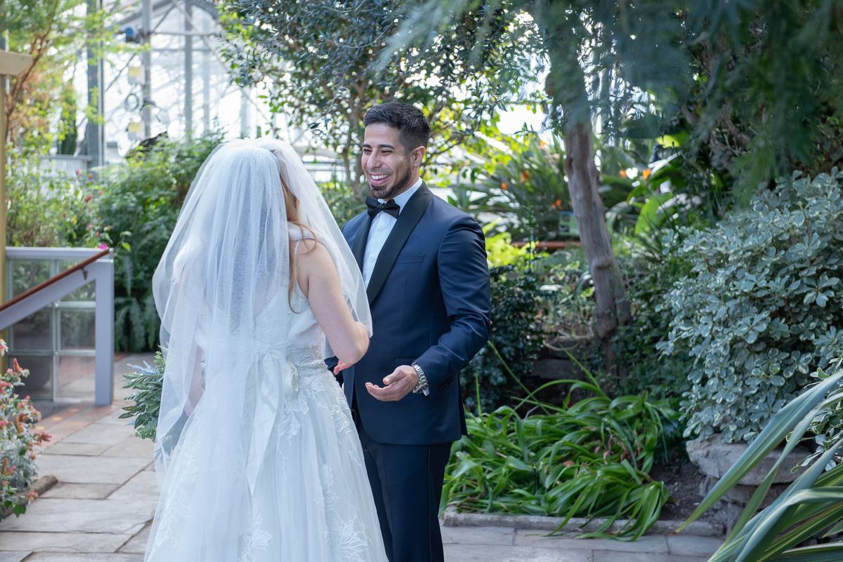 A groom in a navy tuxedo smiles at his bride, whose veil flows behind her, in a greenhouse with lush greenery.