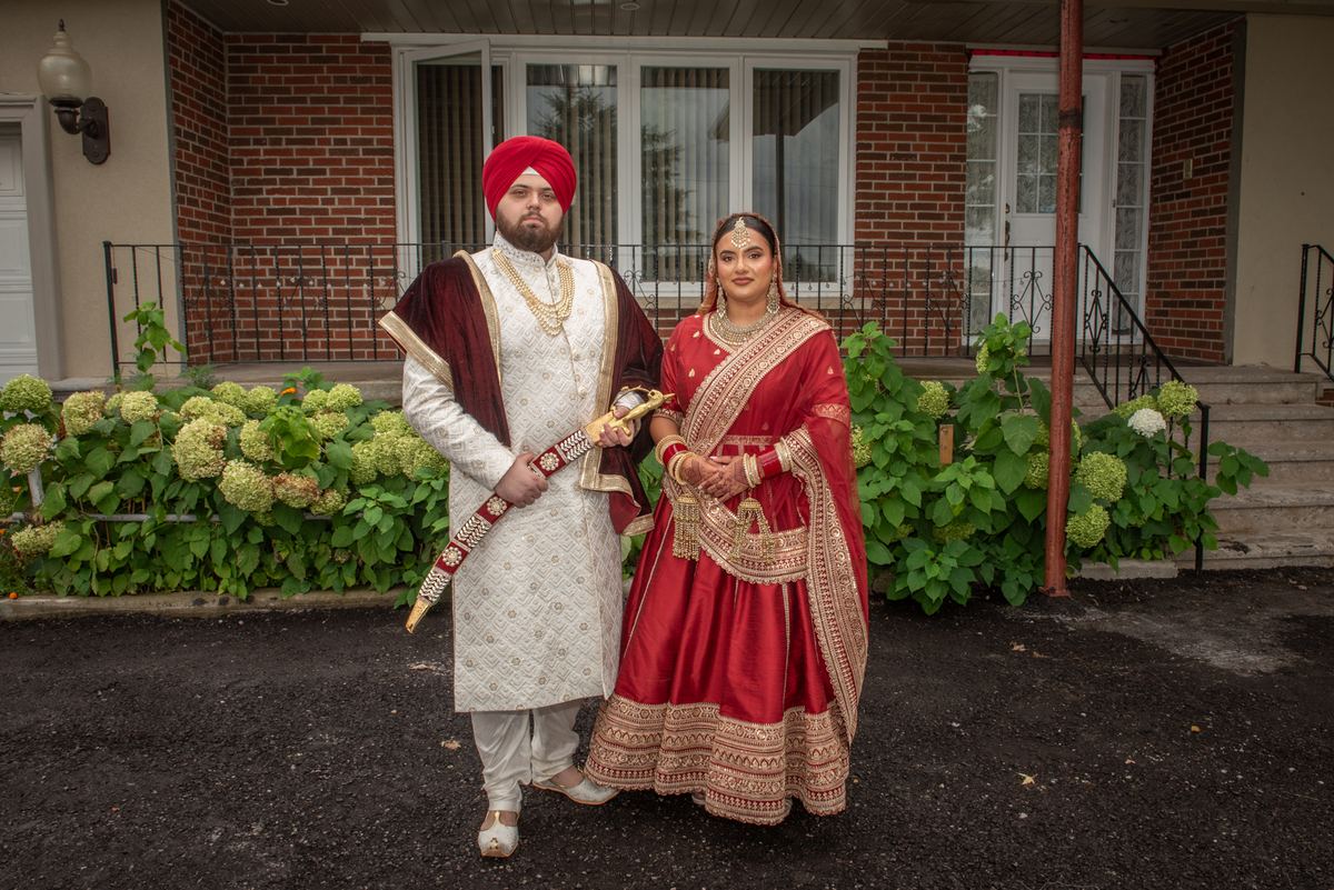 A Sikh newlywed couple poses formally in traditional attire, framed by lush hydrangeas on an overcast day.