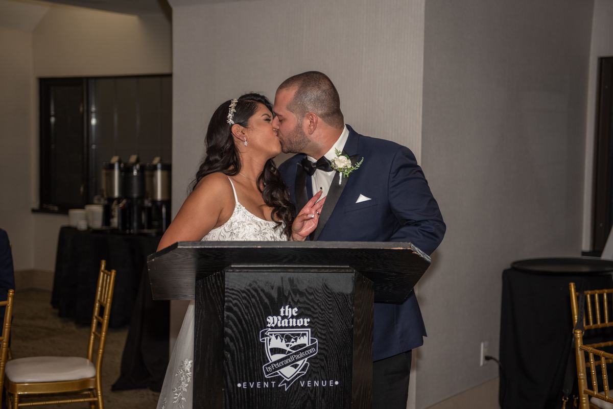A bride and groom exchange a tender kiss at a podium during their reception, surrounded by event chairs and tables.