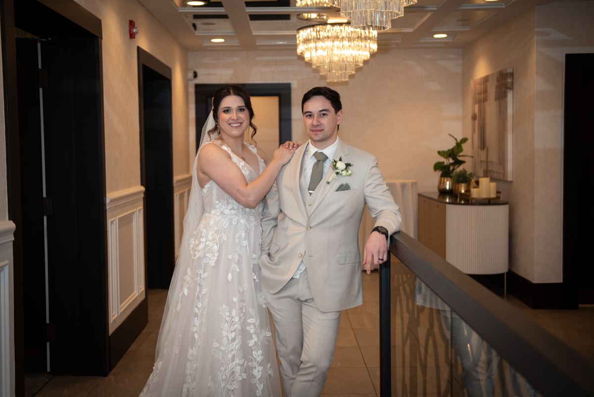 A bride in a white floral gown and groom in a light gray suit smile warmly near a railing under a crystal chandelier.