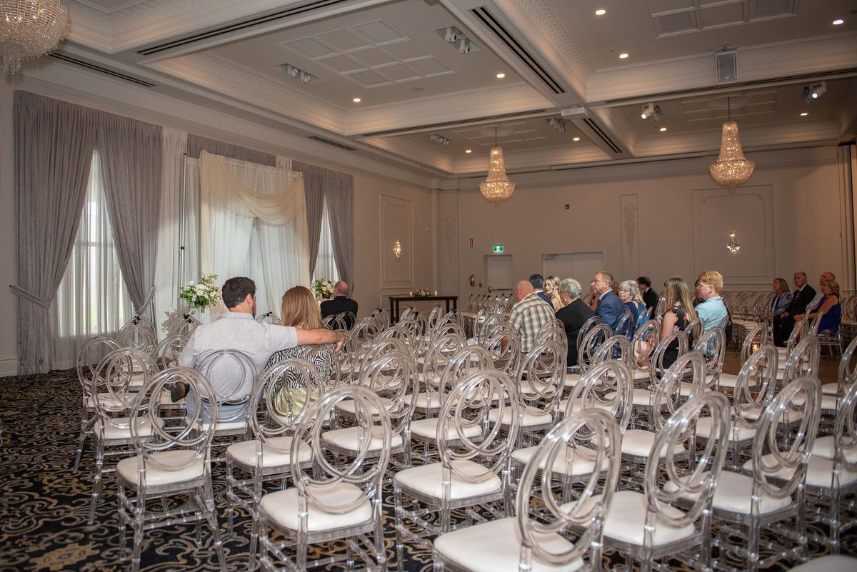 Guests in clear chairs watch a wedding ceremony with natural light and chandeliers illuminating the hall.