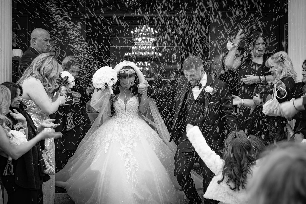 A bride and groom exit amid cheering guests tossing confetti and sparkling lights from an ornate chandelier.