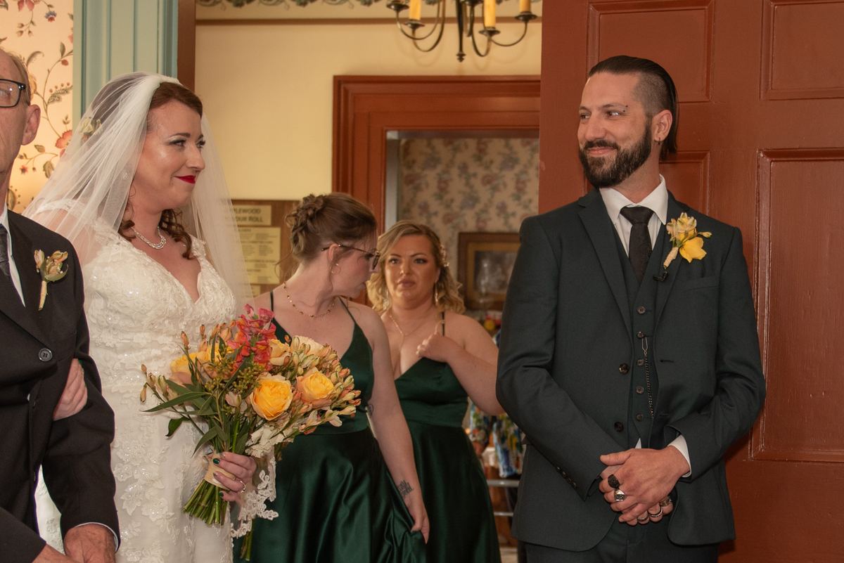 Bride in white dress smiles at groom during ceremony, surrounded by bridesmaids and floral decorations.