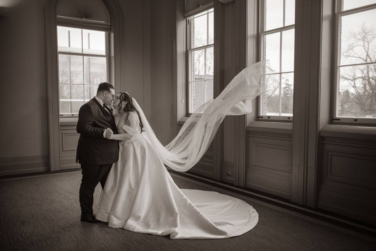 A newlywed couple stands close, gazing at each other in a grand room with streaming natural light and billowing veil.