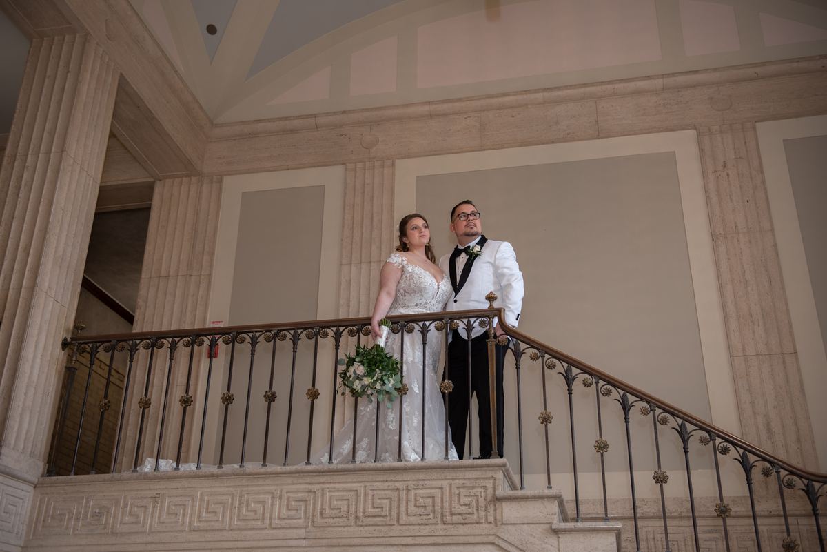 A bride and groom stand on a grand staircase, gazing upward in the softly lit classical space with columns and railing.