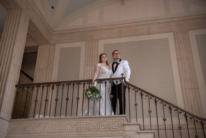 A bride and groom stand on a grand staircase, gazing upward in the softly lit classical space with columns and railing.