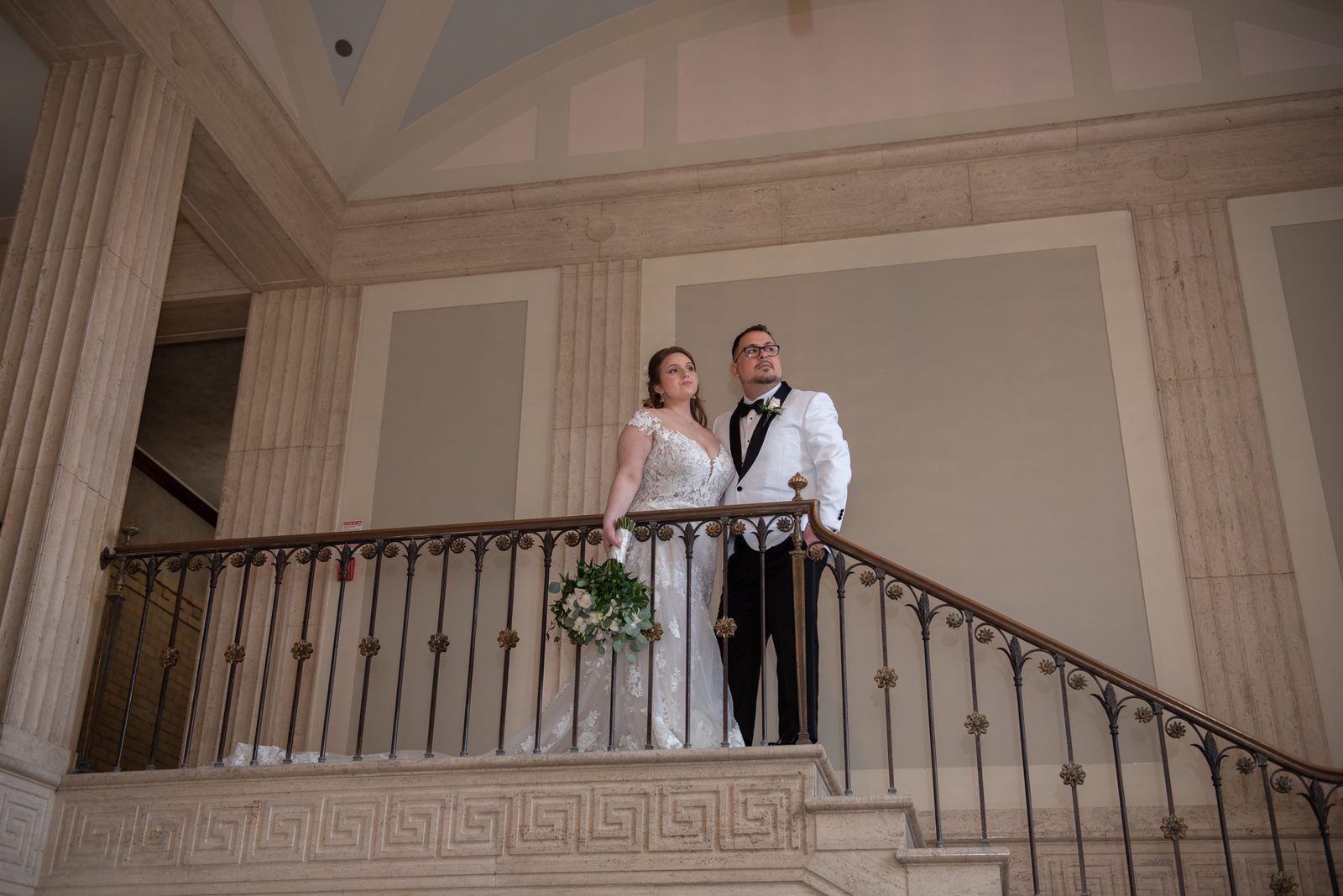 A bride and groom stand on a grand staircase, gazing upward in the softly lit classical space with columns and railing.