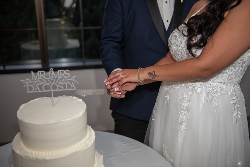 A bride and groom cut their wedding cake together, hands clasped around the knife, with the white frosted cake.