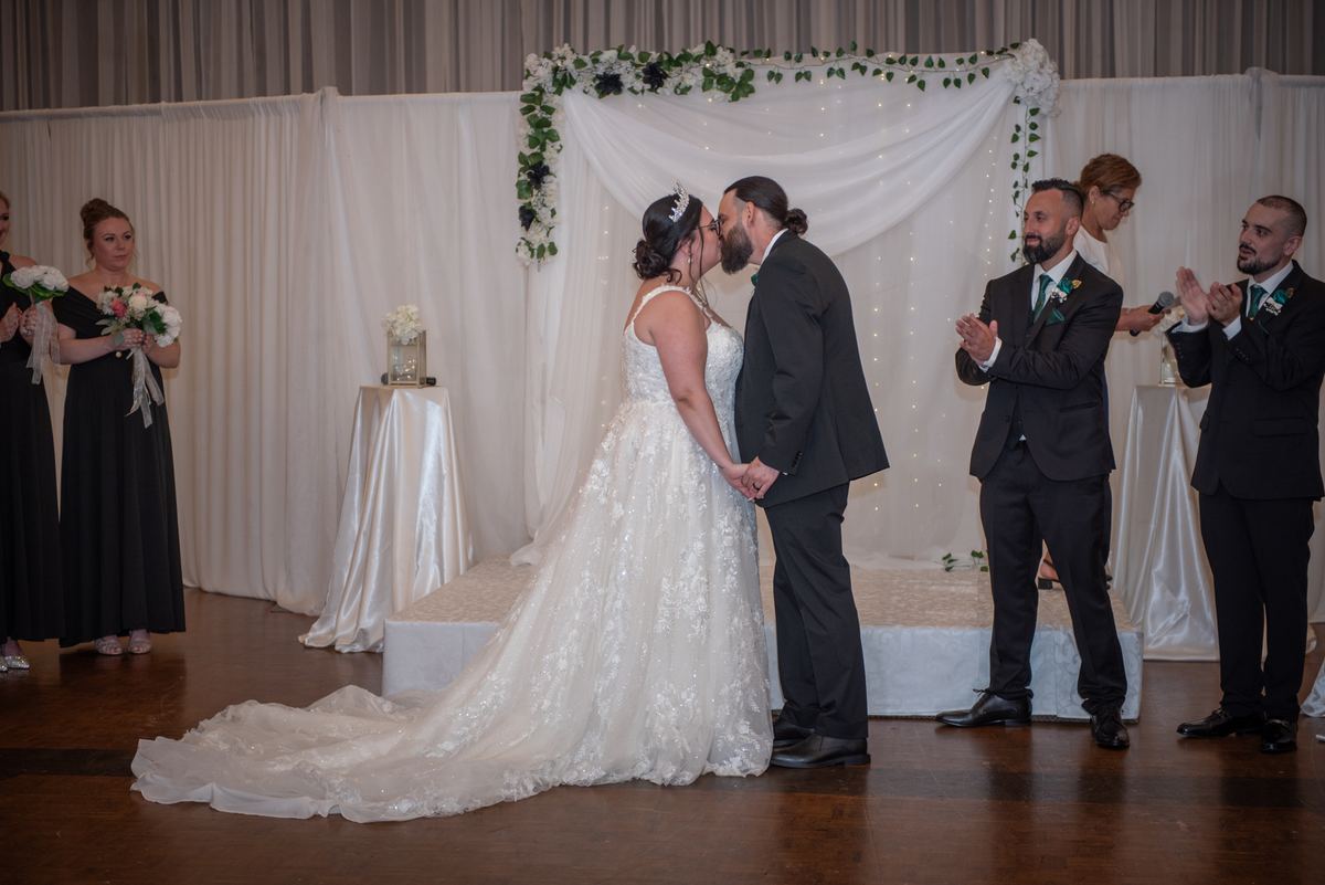 A bride and groom kiss at a wedding ceremony under an arch of white drapery and greenery, with guests applauding.