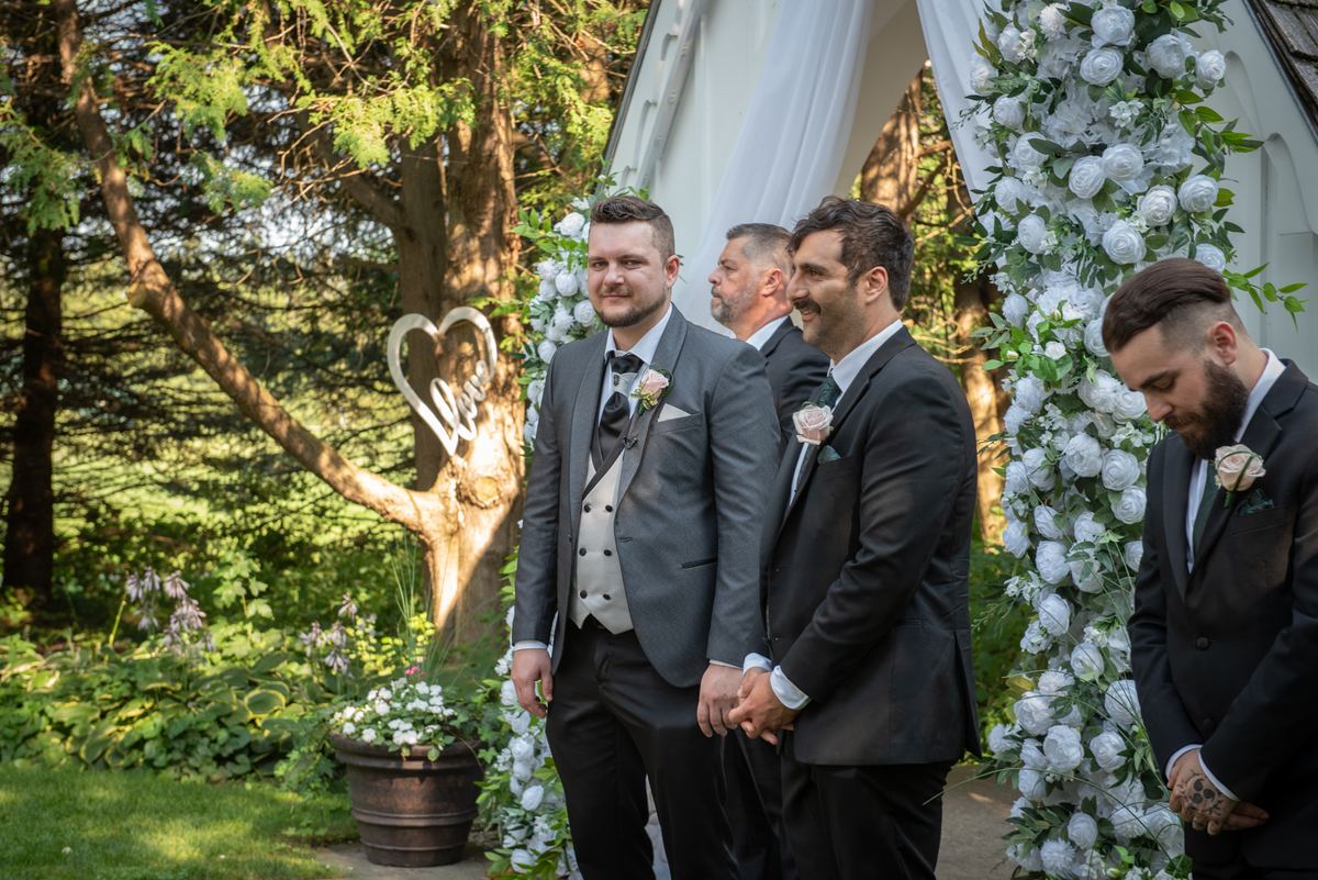 Groomsmen in formal suits stand together, hands clasped, looking toward the ceremony arch under dappled sunlight.