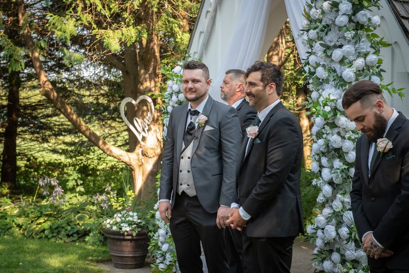 Groomsmen in formal suits stand together, hands clasped, looking toward the ceremony arch under dappled sunlight.