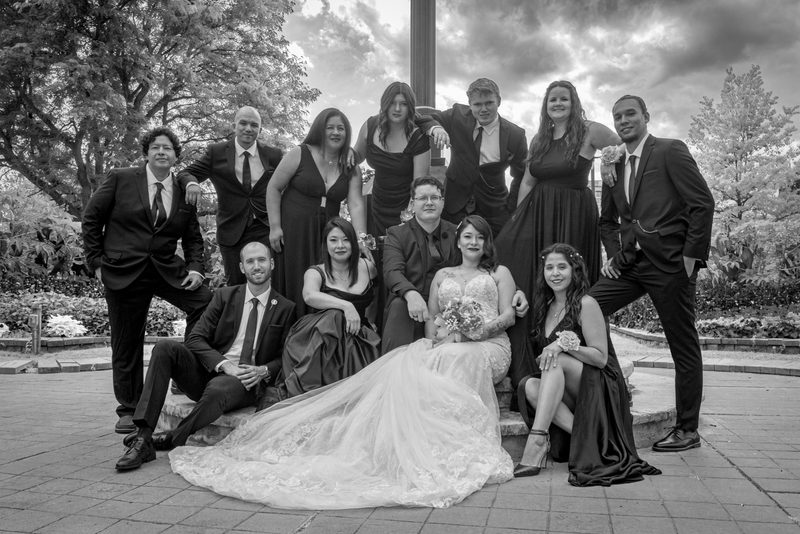 A wedding party poses outdoors on a terrace under a cloudy sky, with the bride in a lace gown front.