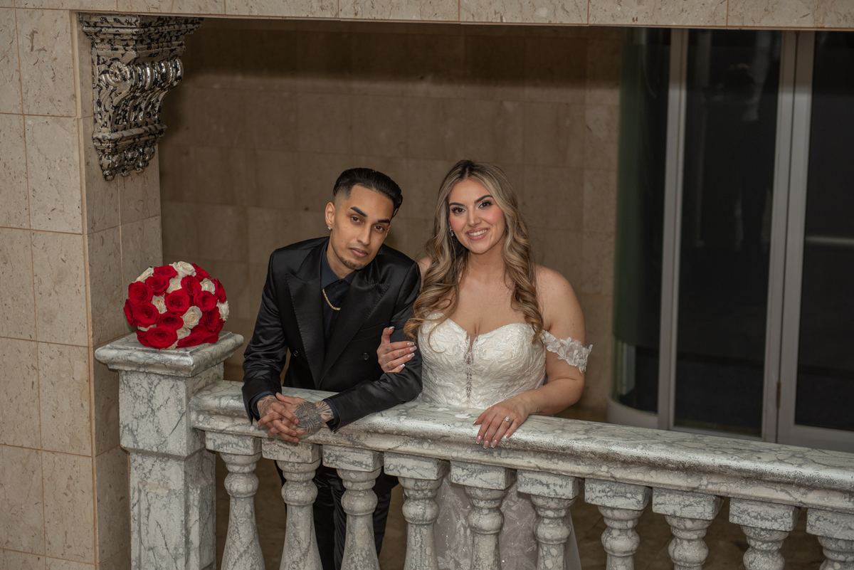 A newlywed couple in formal attire leans on a marble balcony railing, with a red-and-white rose bouquet nearby.