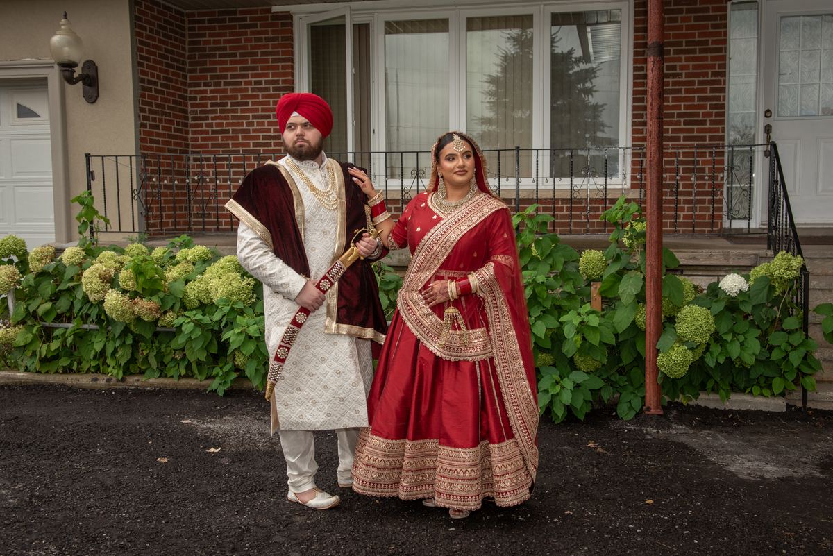A Sikh groom in white and red with sword stands beside his bride in a red and gold lehenga by a brick house.