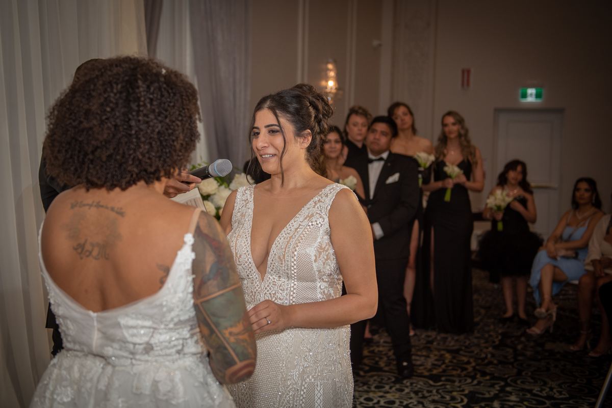 A bride with tattoos speaks into a microphone to her smiling partner in front of formal-clad guests.