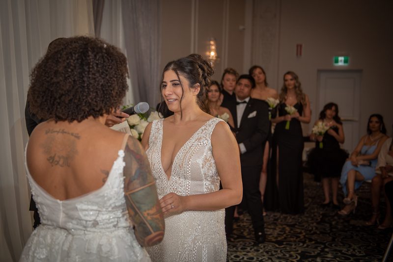 A bride with tattoos speaks into a microphone to her smiling partner in front of formal-clad guests.