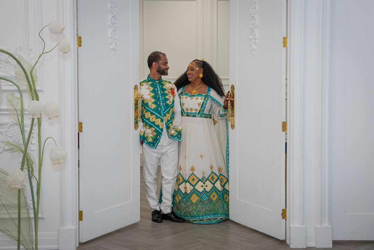 A smiling couple in traditional attire stands by a door, hands touching, in a bright, ornate setting.