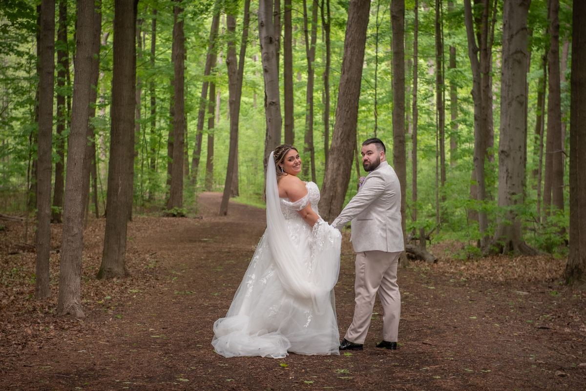 Bride in white dress and veil smiles over shoulder as groom in light suit holds her hand on forest path.