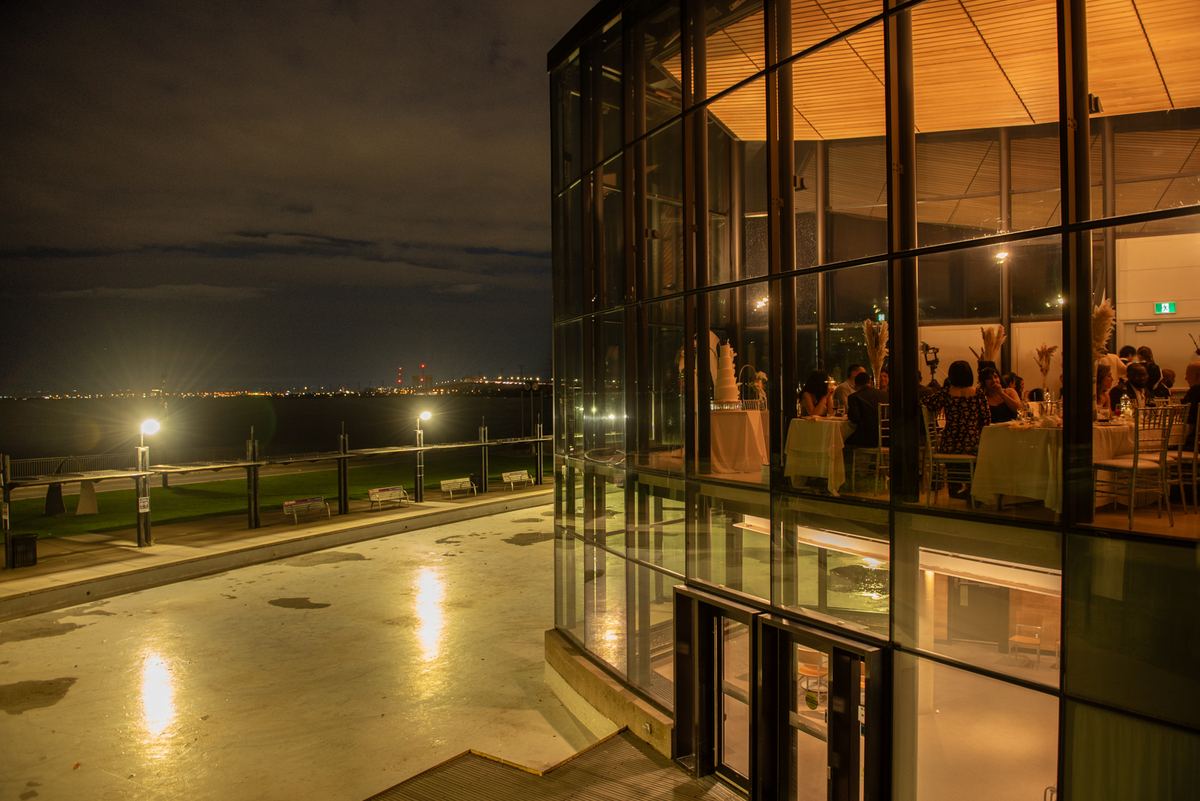 Guests celebrate at a modern glass-walled reception with a waterfront view, reflecting city lights and a distant skyline.