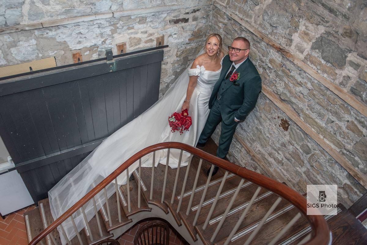 Bride in white gown and groom in dark pinstripe suit smile on wooden staircase inside rustic stone building.