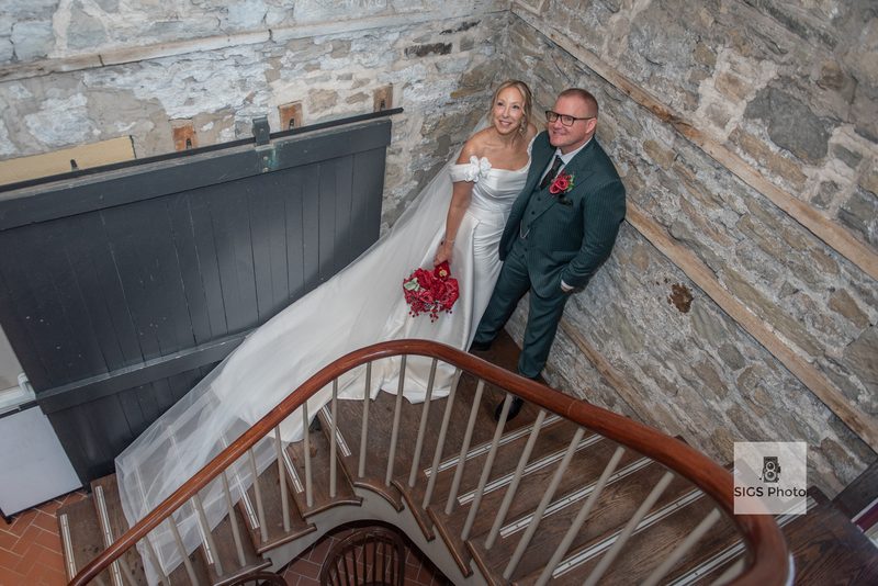 Bride in white gown and groom in dark pinstripe suit smile on wooden staircase inside rustic stone building.