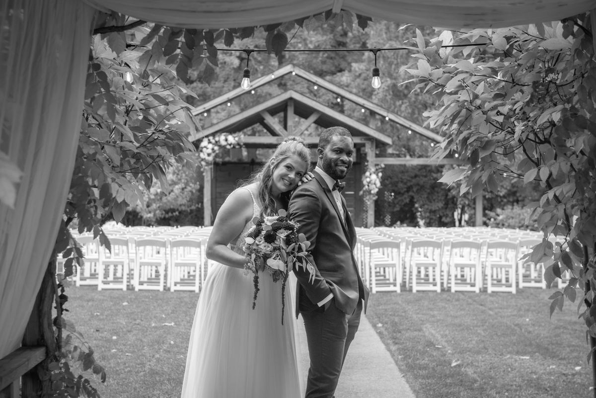 A smiling bride and groom stand close under a leafy archway at night, surrounded by string lights and white chairs.
