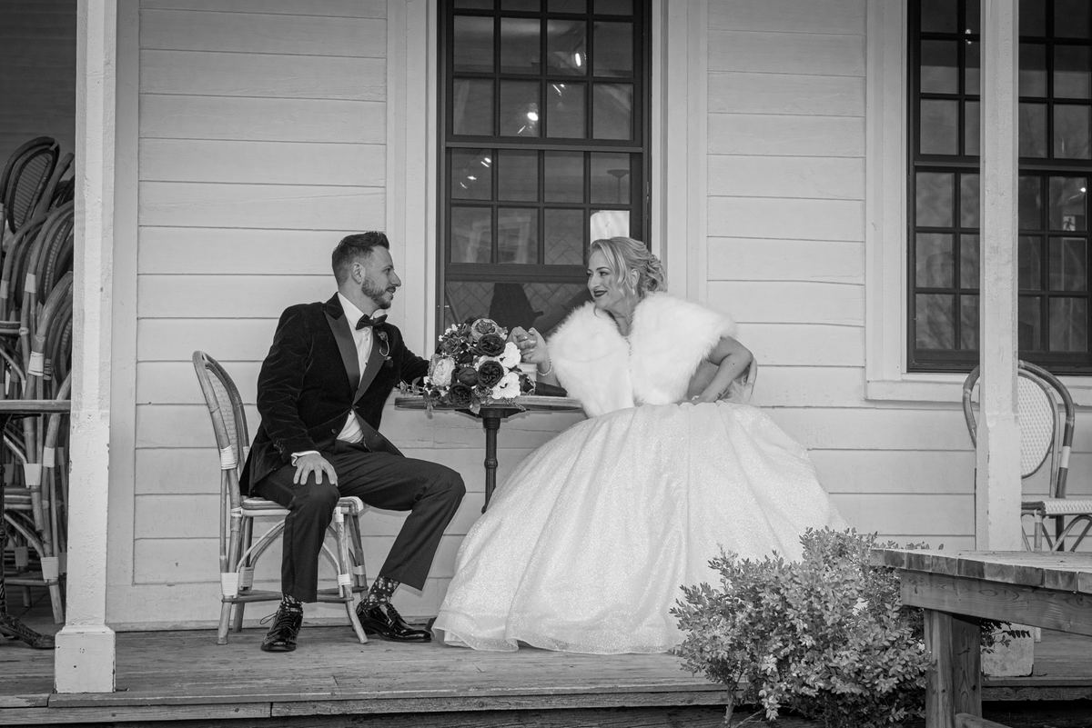 A newlywed couple smiles at each other across a table with flowers on a rustic porch in soft daylight.