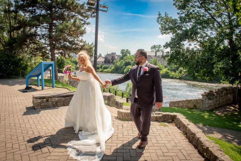 A bride and groom walk hand-in-hand along a stone path by the river, smiling as they pass blue buildings and trees.