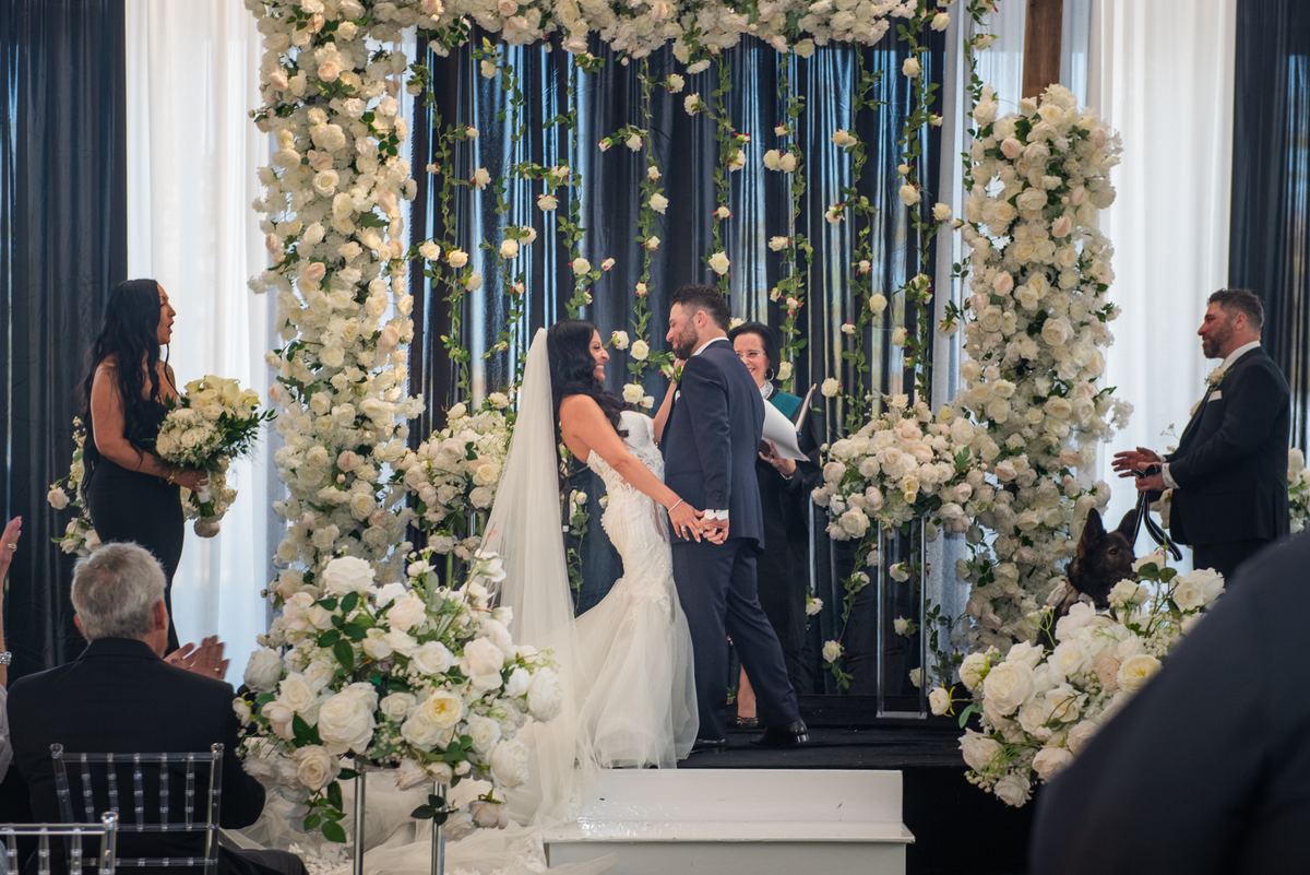 Bride and groom stand hand-in-hand on a floral-draped stage during their ceremony, surrounded by guests.