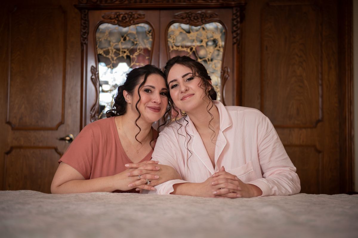 Two women lean together with gentle smiles, resting foreheads on a bed under soft stained-glass light.