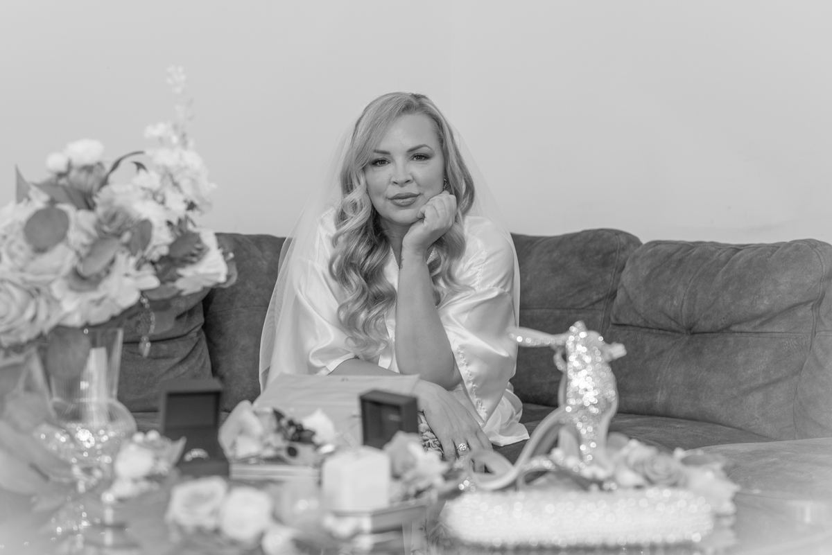 A bride sits thoughtfully on a couch surrounded by wedding decor and flowers in soft black-and-white tones.