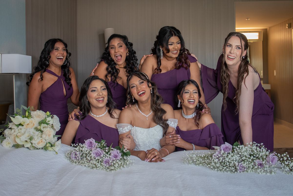 A bridal party laughs and poses around the bride on a bed adorned with lavender and white flowers, filled with joy.