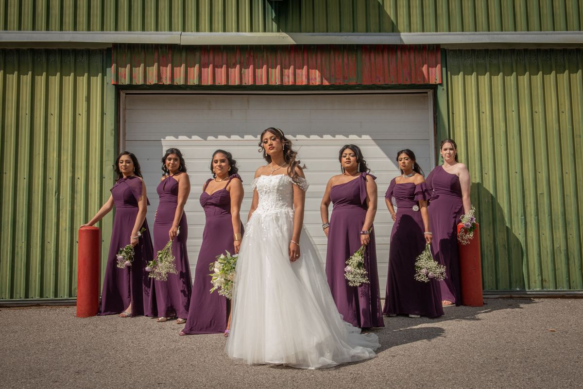 A bride in white stands with her purple-clad bridesmaids before an industrial building, sunlight casting sharp shadows.