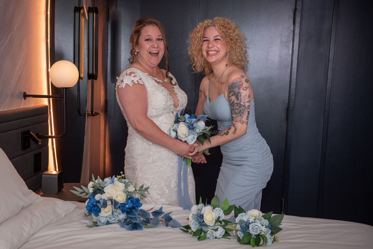 Two brides laughing with a shared bouquet near a bed decorated with floral arrangements and warm lighting from a wall sconce.