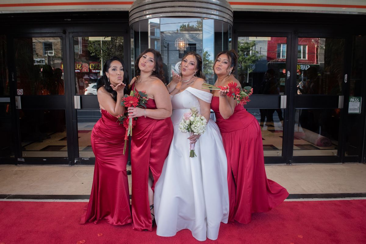 Four women, including a bride in white, blow kisses while holding bouquets on a red carpet outdoors.